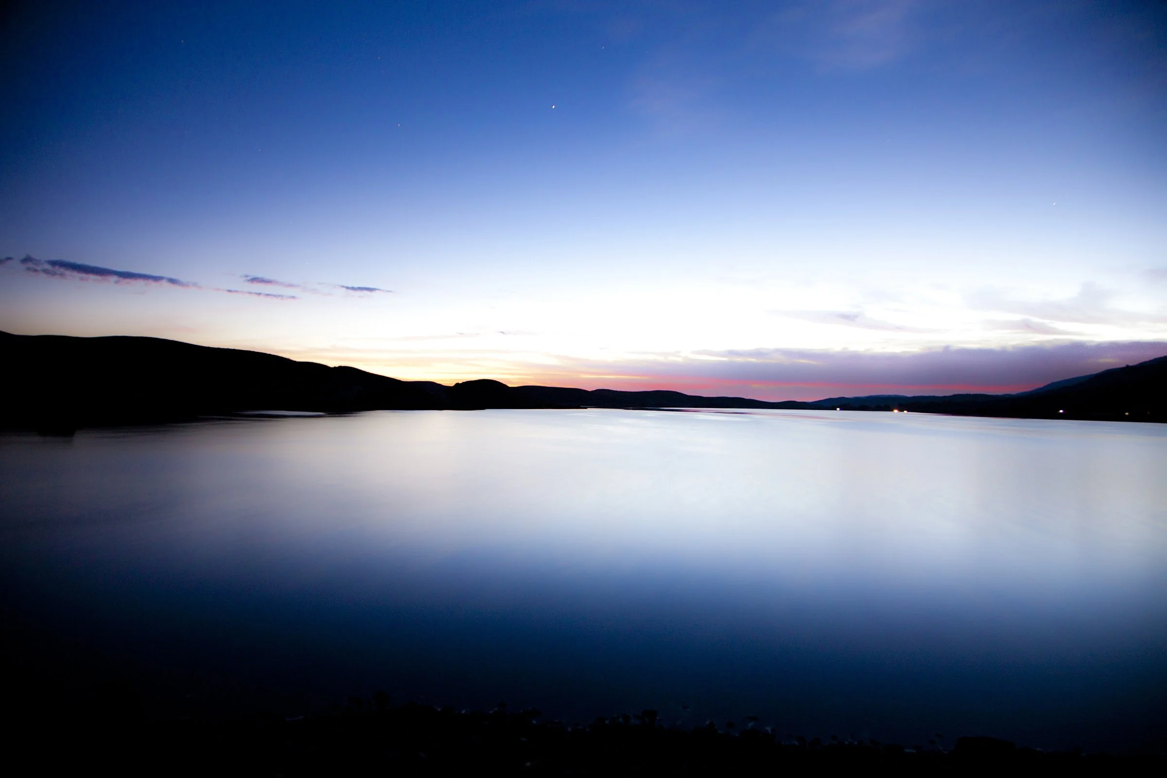 A scenic view of a calm lake at sunset with a silhouette of hills in the background and a sky transitioning from light to dark, with a few clouds and stars beginning to appear.