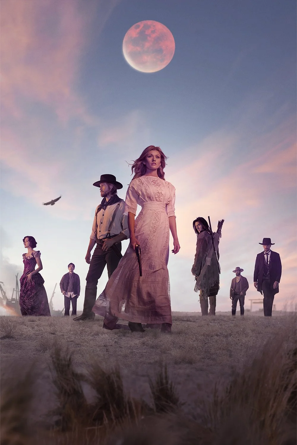Group of people standing outdoors at sunset with a pink moon in the sky, some holding guns and arrows, dressed in vintage and rugged clothing.