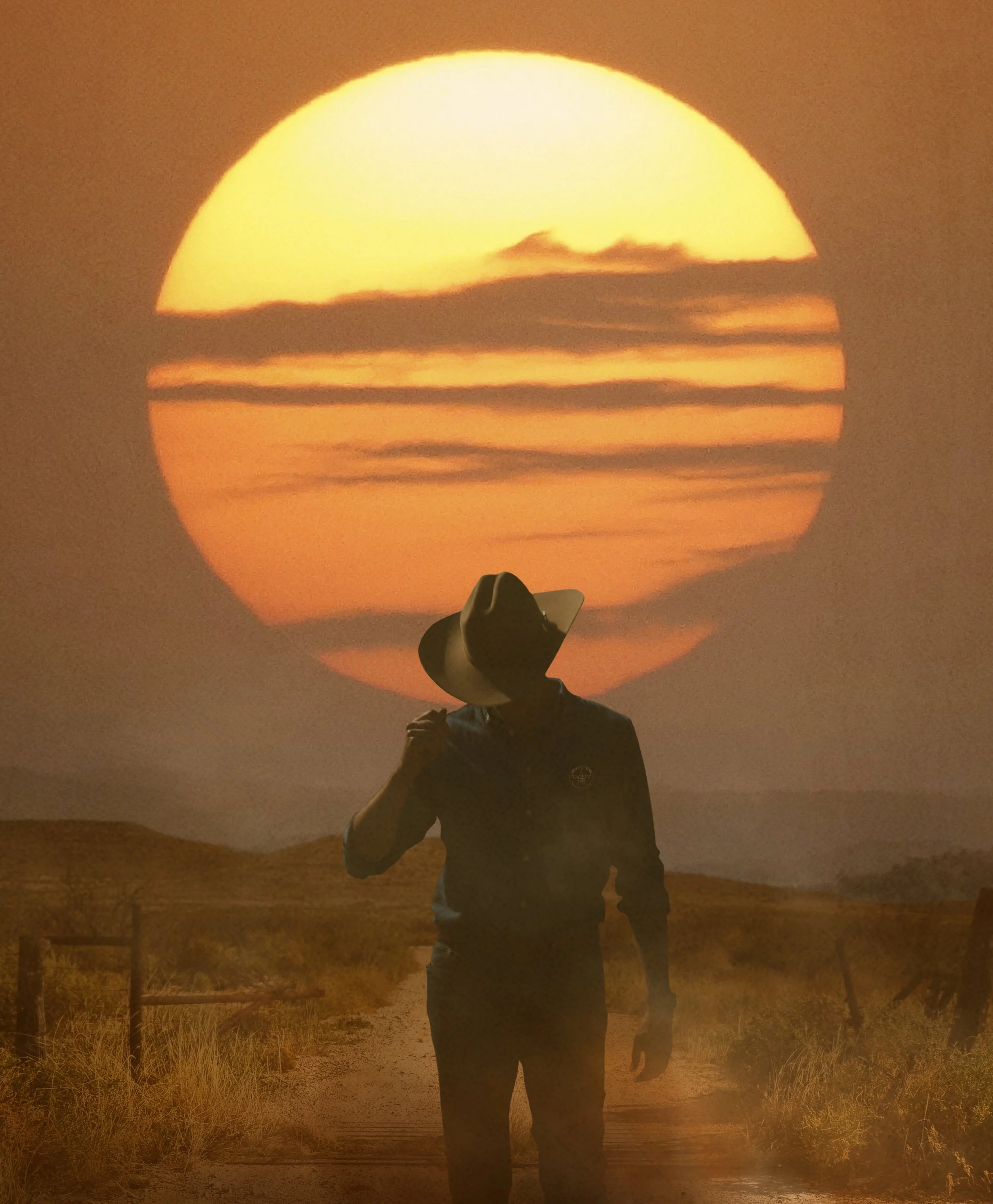 A person standing in a field at sunset with the sun appearing as a large, bright, orange sphere, partially obscured by clouds, with a hat and dark clothing.