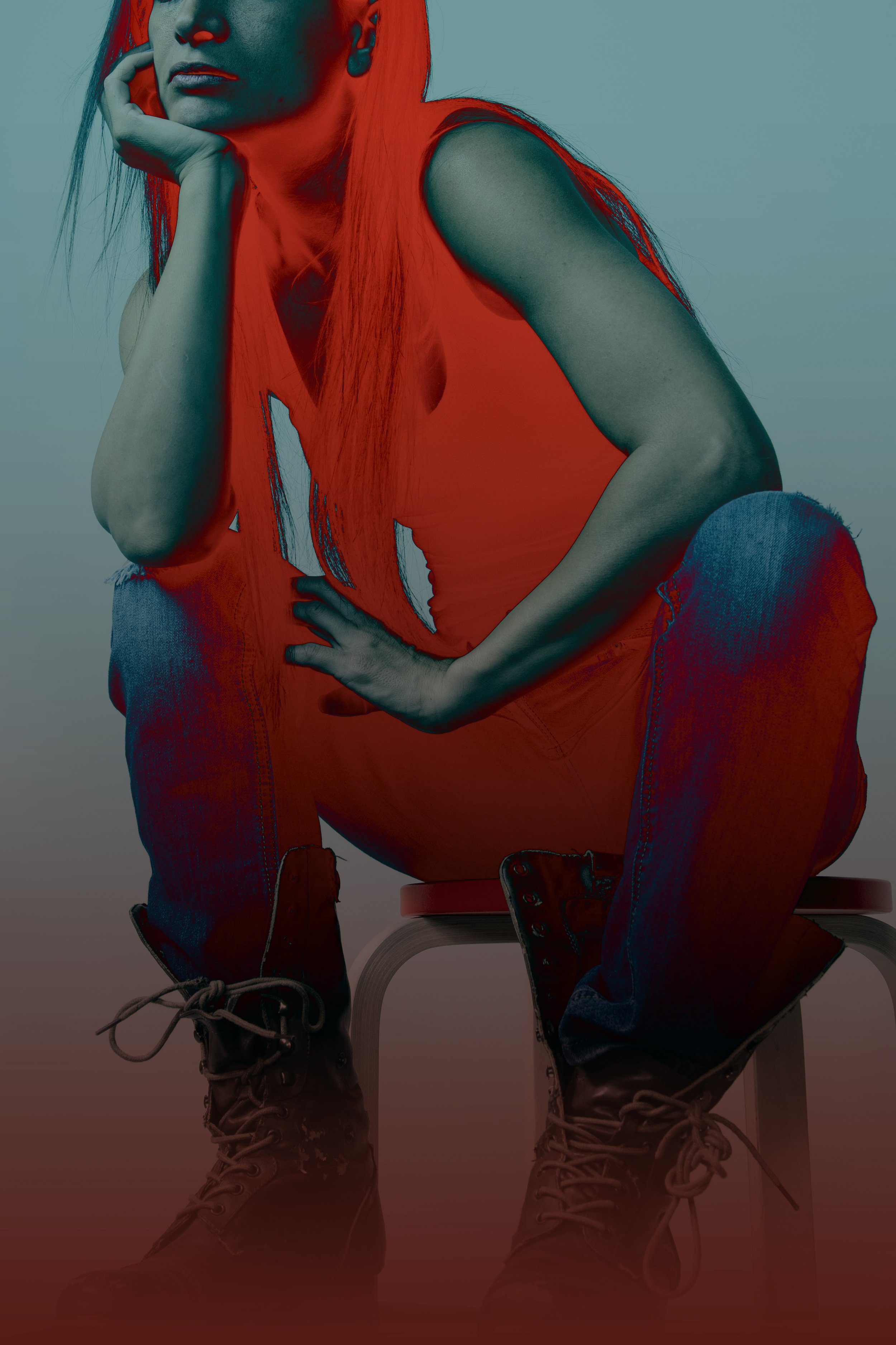 A person with long hair wearing a sleeveless orange top, jeans, and boots, sitting on a stool with one hand resting on their face, in a studio with a plain background.