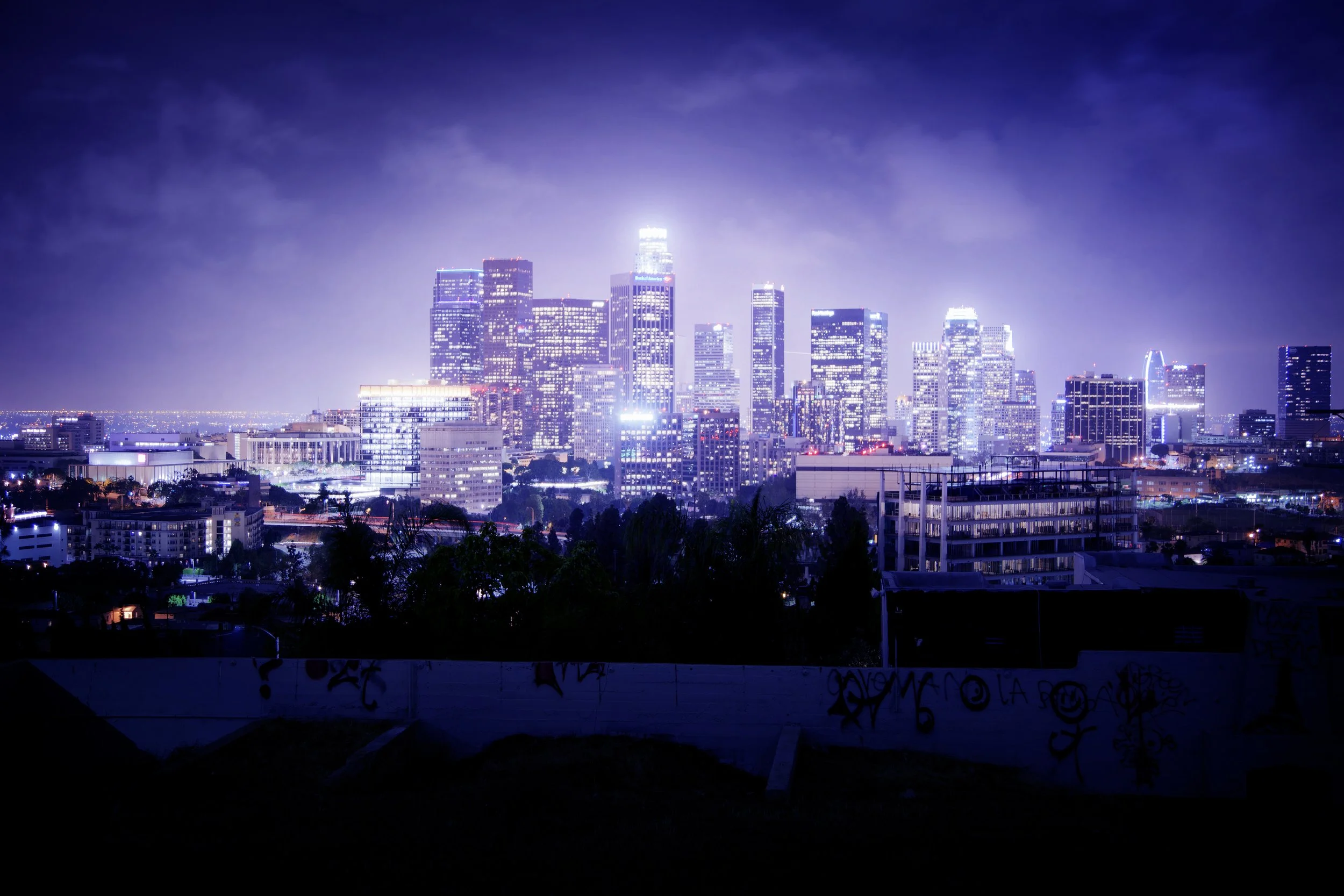Night city skyline with illuminated skyscrapers under a cloudy purple sky, viewed from a distance.