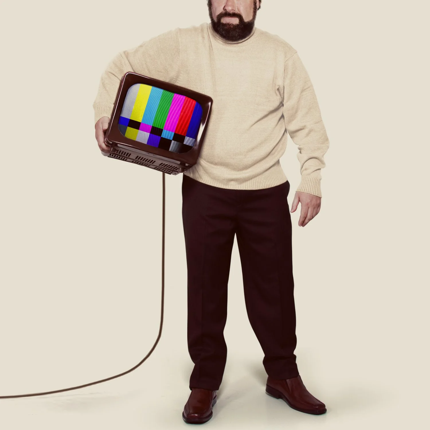 A man in a beige sweater and dark pants holding a vintage television with a colorful test pattern, with a beige background.