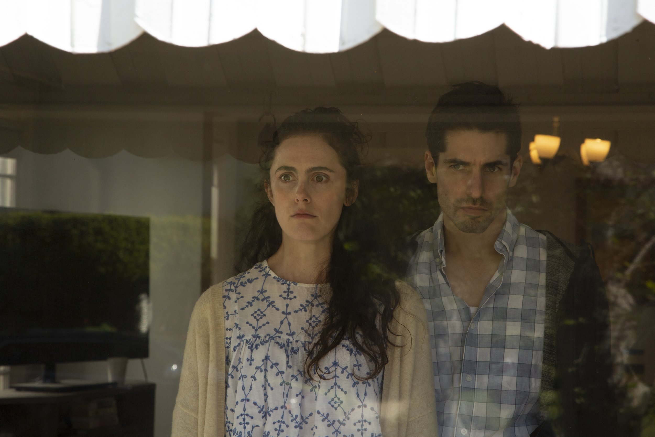A woman and a man looking through a window, their reflection visible. The woman has dark curly hair and is wearing a light-colored cardigan over a patterned blouse. The man has short dark hair and is wearing a checked shirt.