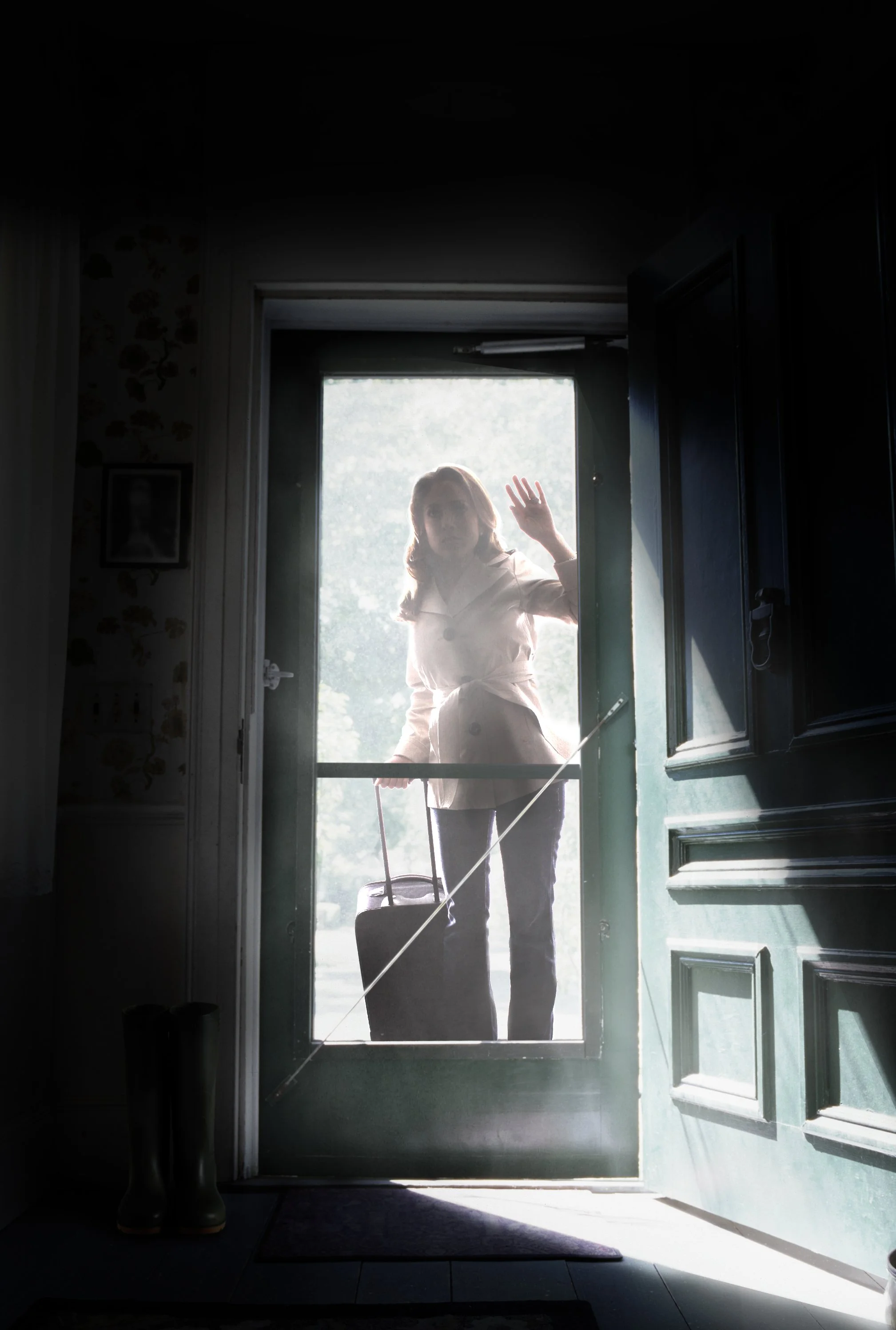 A woman standing outside a door, waving, with a rolling suitcase, illuminated by sunlight behind her.