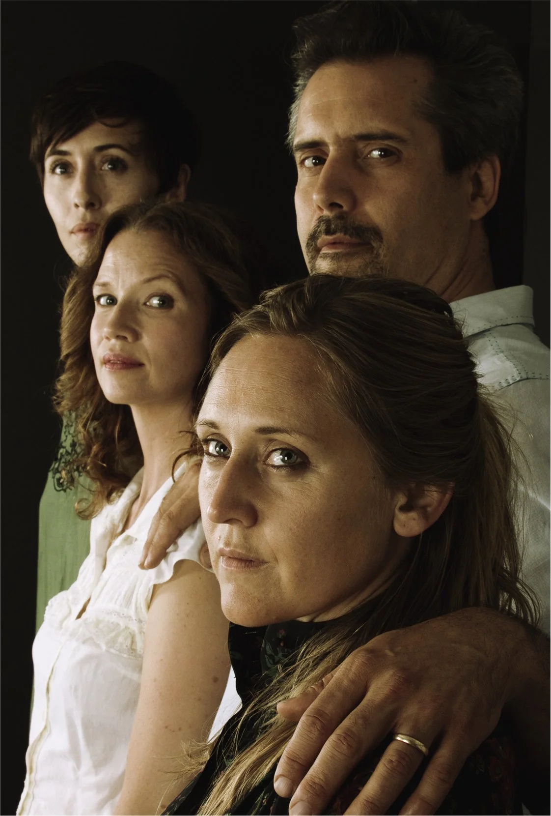 Group portrait of four adults, two women and two men, posing closely against a dark background. The woman in front has red hair and blue eyes, and the woman behind her has curly brown hair. One man has gray hair and a mustache, and the other woman ha