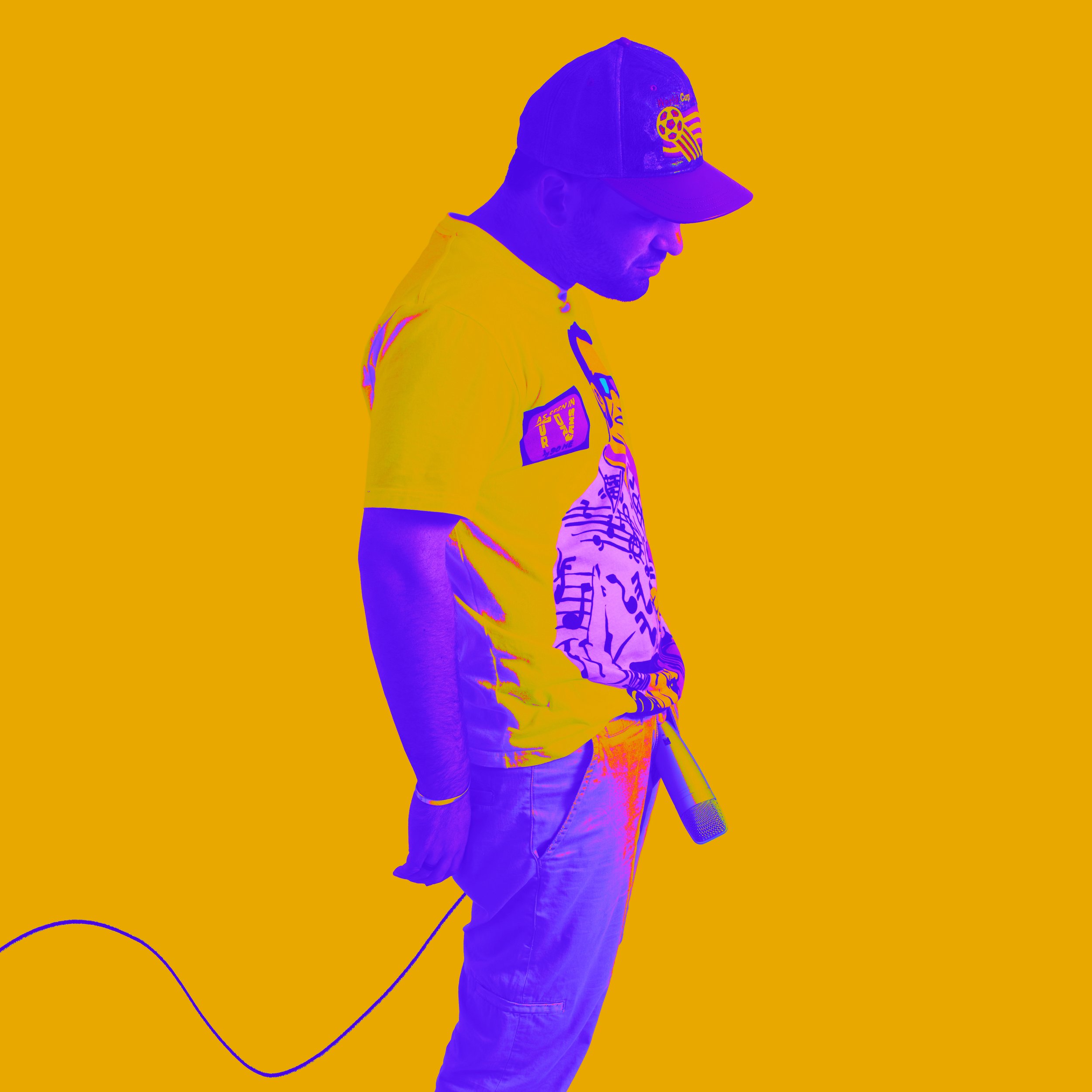 A person wearing a baseball cap and a yellow shirt with printed patches, holding a microphone connected to a coiled cable, standing against an orange background.