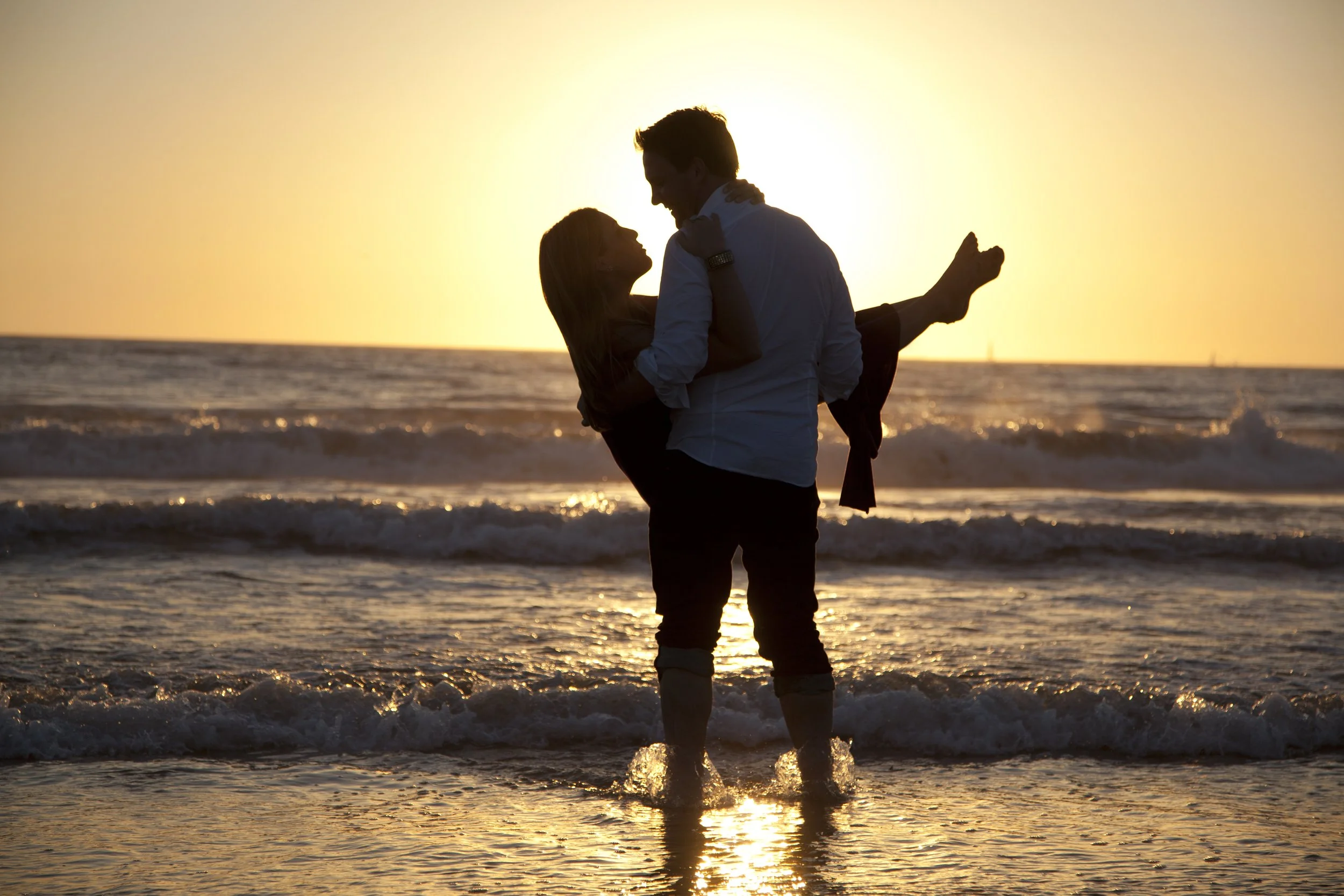 A couple standing in the ocean at sunset, with the man holding the woman in his arms, smiling and looking at each other.