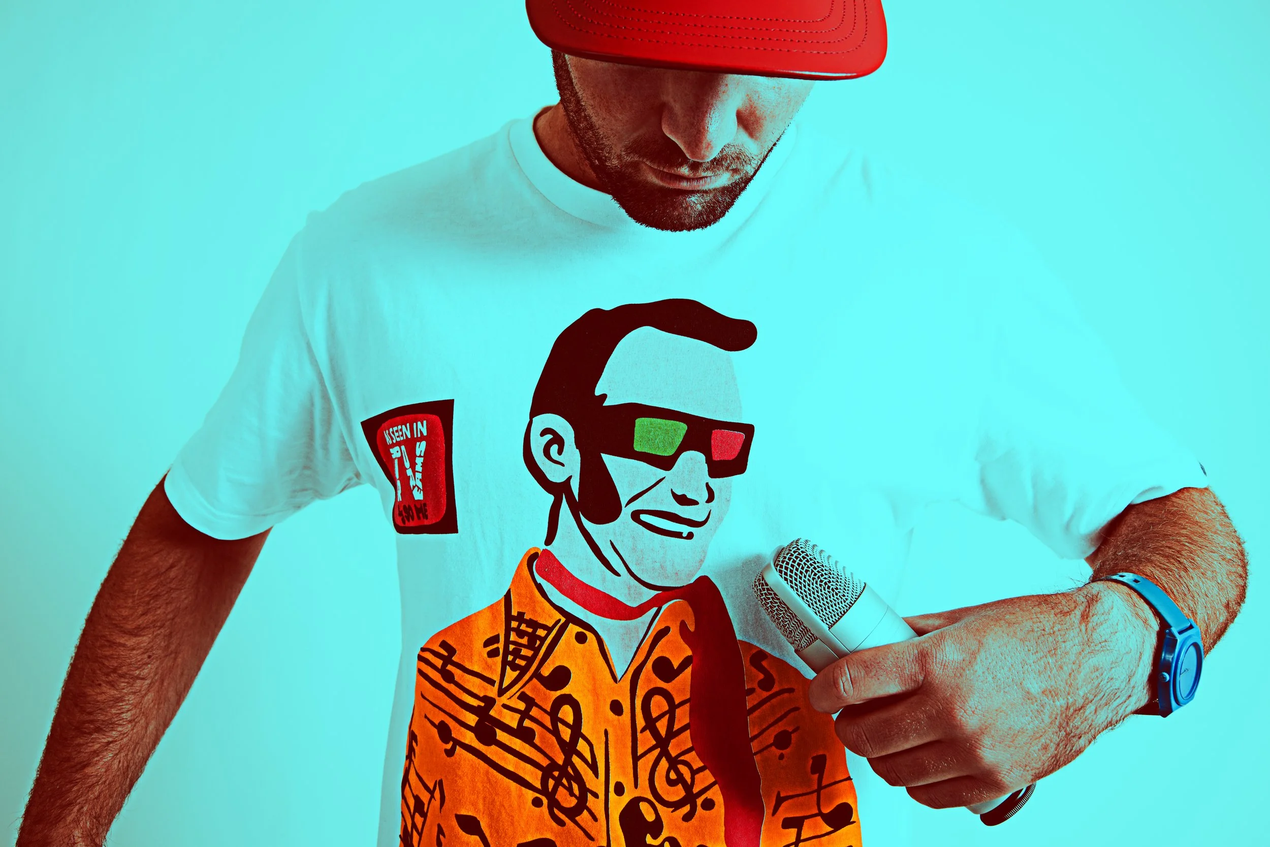 A man wearing a white t-shirt with a colorful graphic of a cartoon character and a red hat, holding a microphone, with a turquoise background.