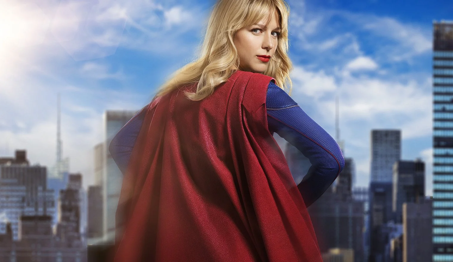 A woman dressed as a superhero stands on a rooftop in a city skyline during daytime, wearing a red cape and a blue suit with textured sleeves.