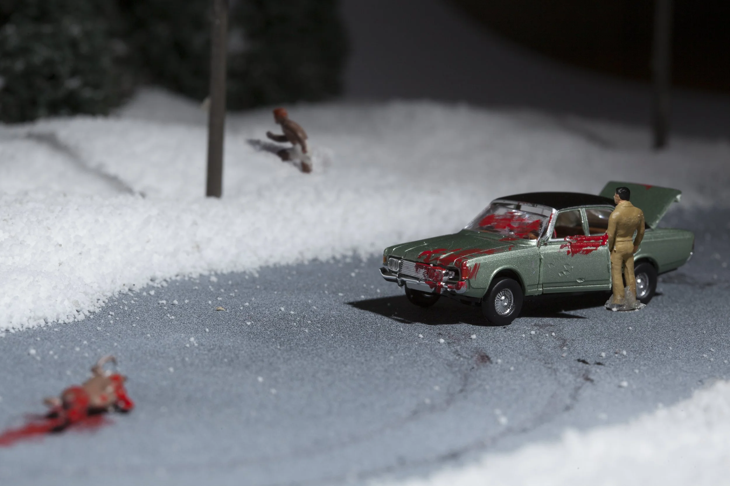 Miniature scene of a green toy car with red splatters, a figure standing outside the car, and small figurines in the snow. The background includes snow-covered ground, trees, and presents.
