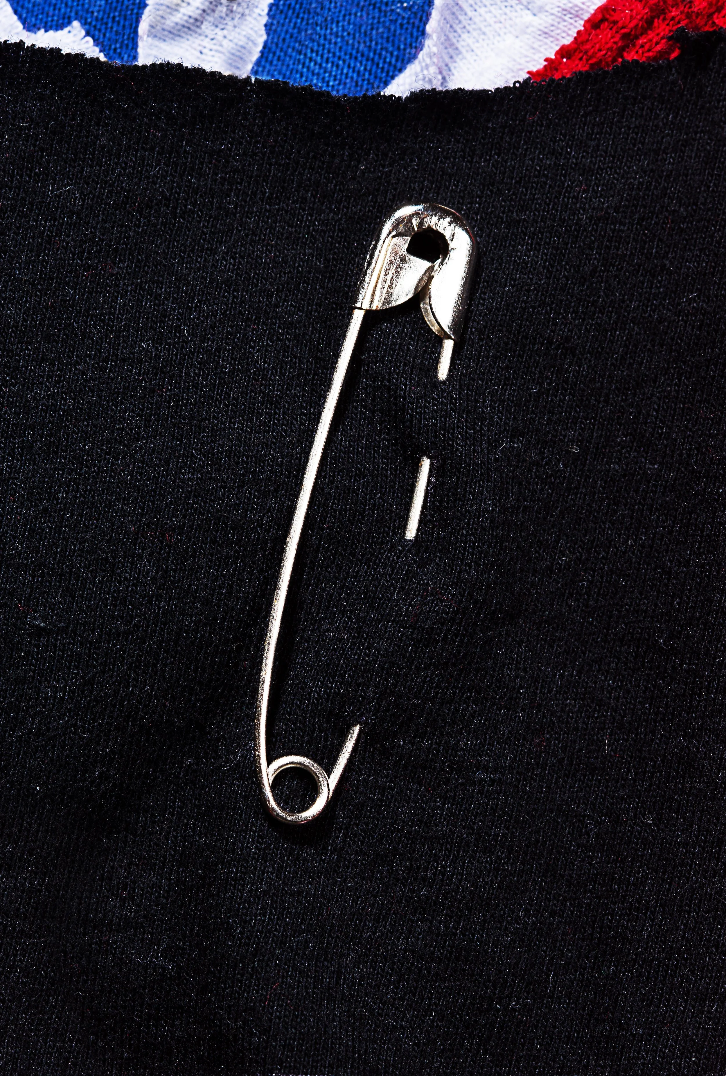 Close-up of a large safety pin attached to black fabric, with some part of colorful clothing visible at the top.