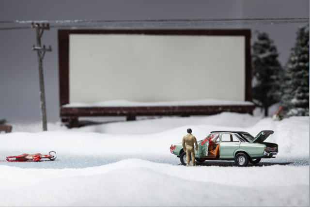 Miniature scene of a soldier figure near an open car trunk in a snowy landscape with a billboard in the background.