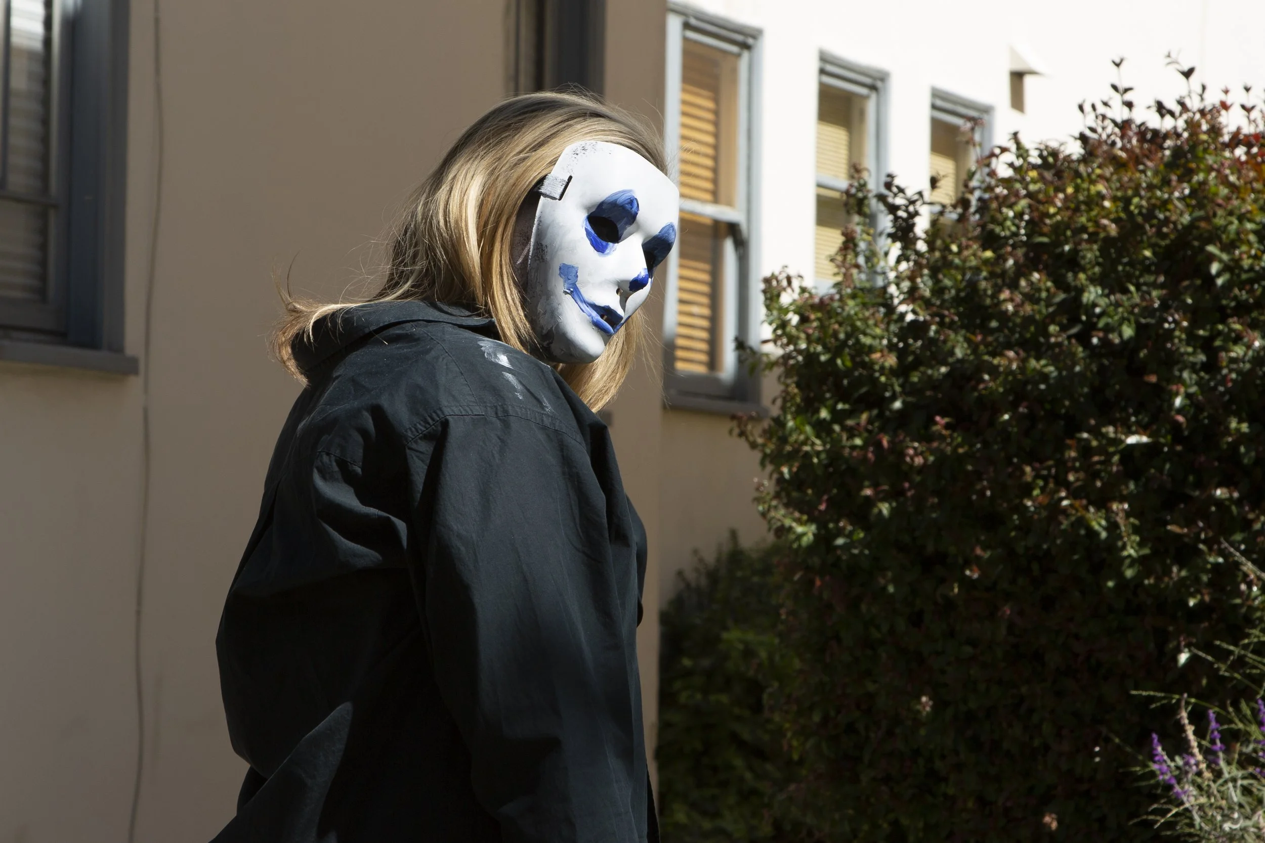Person wearing a white skull mask with blue accents, standing outdoors near a building with windows and bushes.