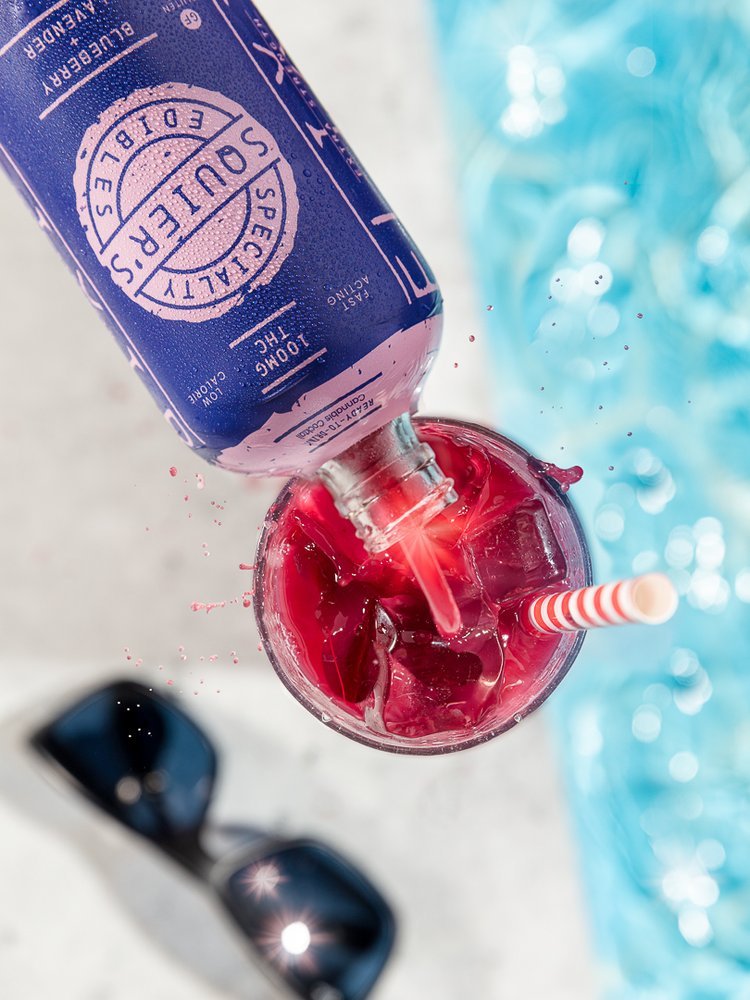 Overhead view of a bottle pouring red liquid into a glass with ice and a striped straw, beside sunglasses and a pool.