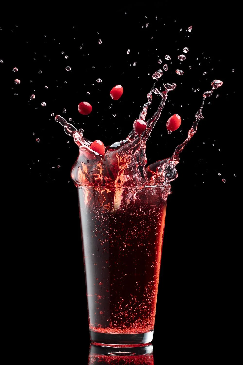 A clear glass of red fizzy liquid with cranberries splashing and bubbles on a black background.