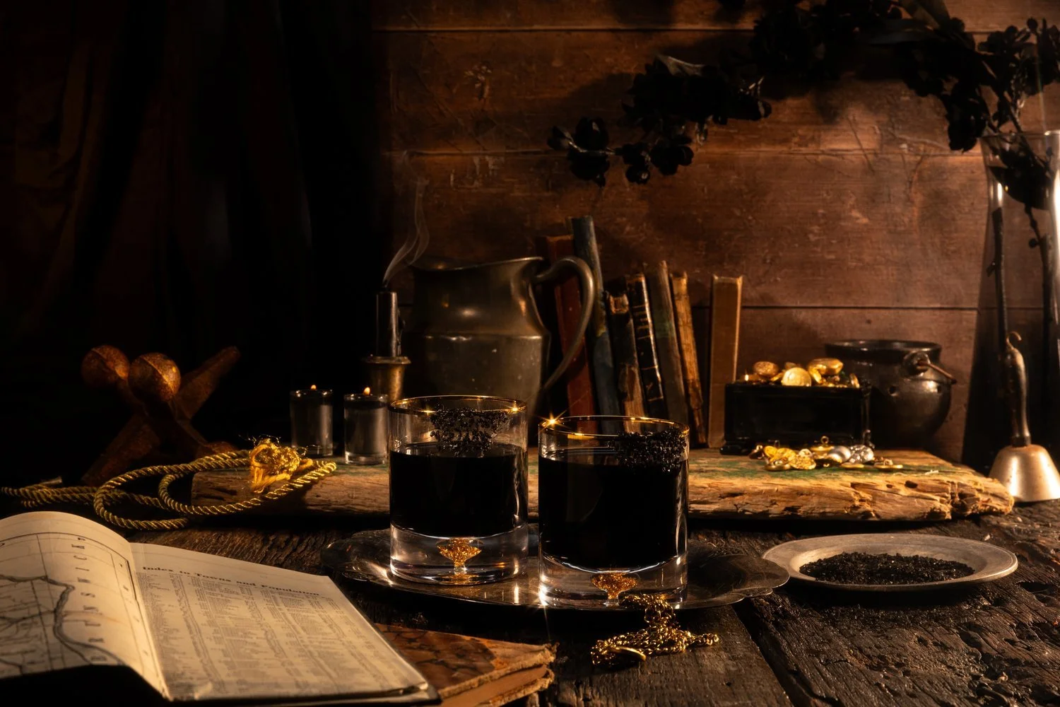 Vintage pirate-inspired still life with two glasses of dark liquid, a smoking candlestick, books, a map, gold coins, and a rope on a wooden table.