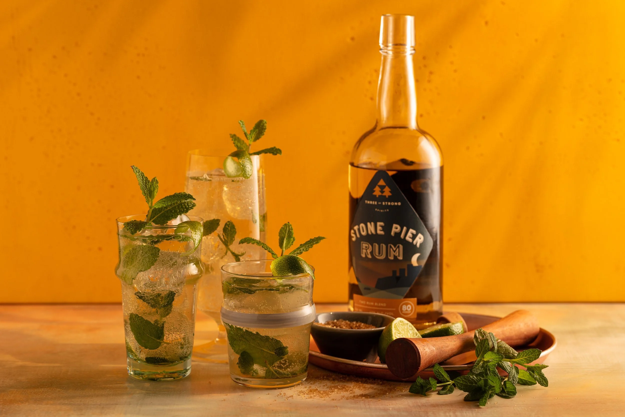 Three glasses of mojito with mint and lime, a bottle of Stone Pier rum, cut lime, brown sugar in a bowl, and a wooden muddler on a textured surface with an orange background.
