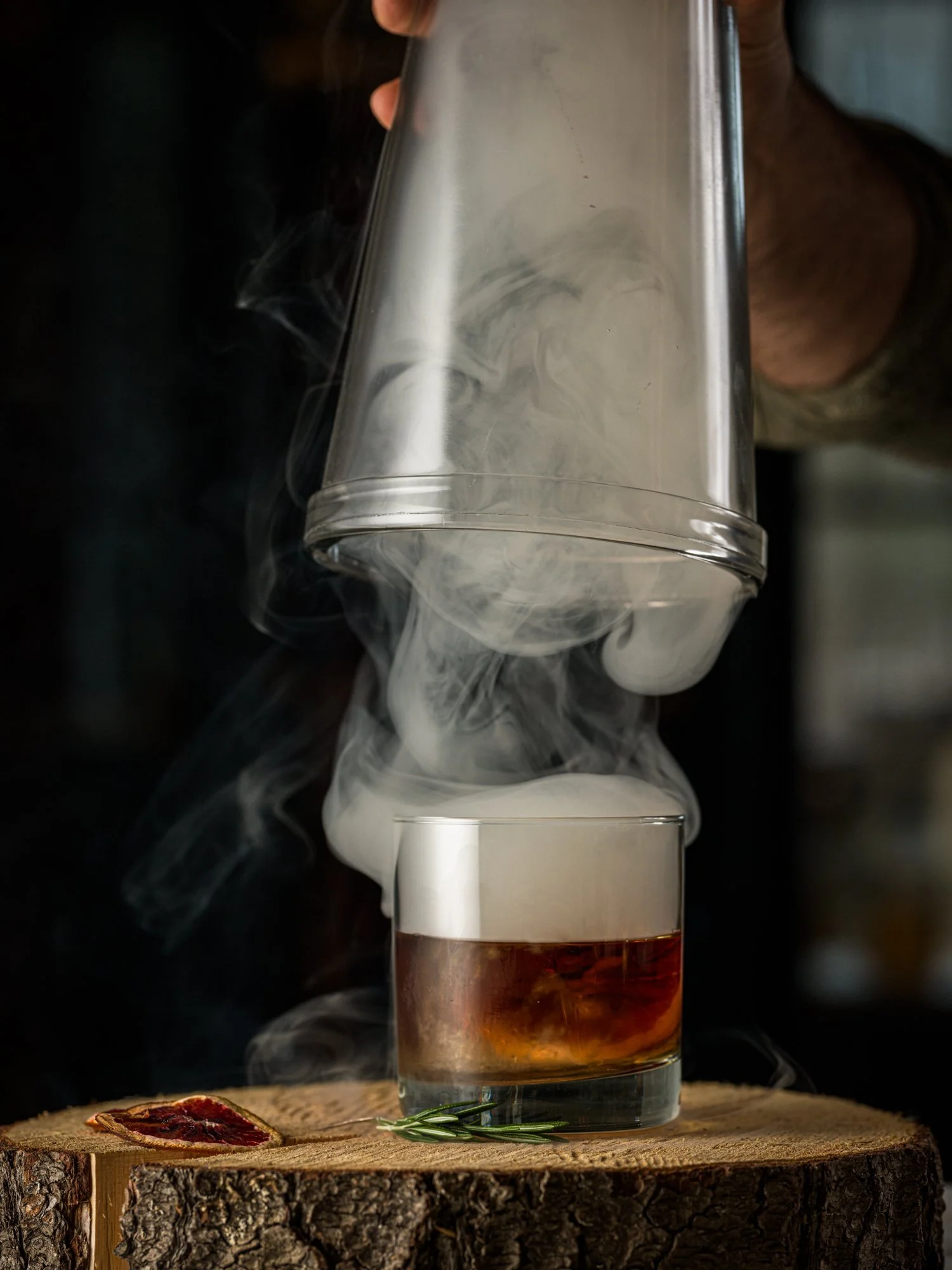 A hand lifting a smoking glass dome from a cocktail in a short glass on a wooden surface, with rosemary and a dried citrus slice nearby.