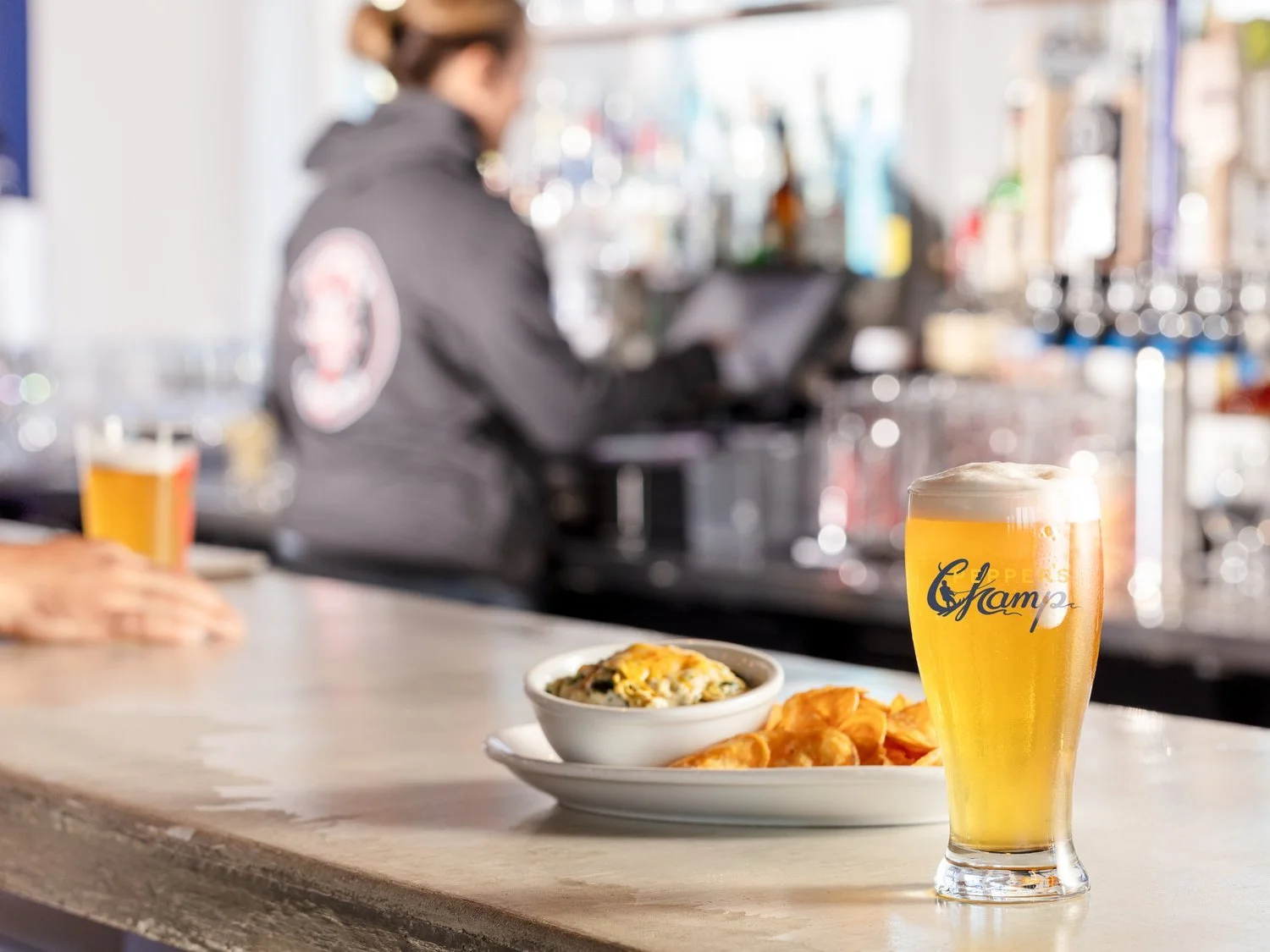 Glass of beer and a plate with dip and chips on a bar counter, with a bartender in the background.