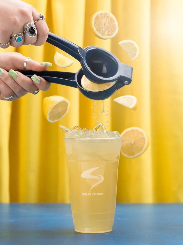 Hand squeezing lemon into a cup of lemonade with ice, surrounded by floating lemon slices, over a yellow background.