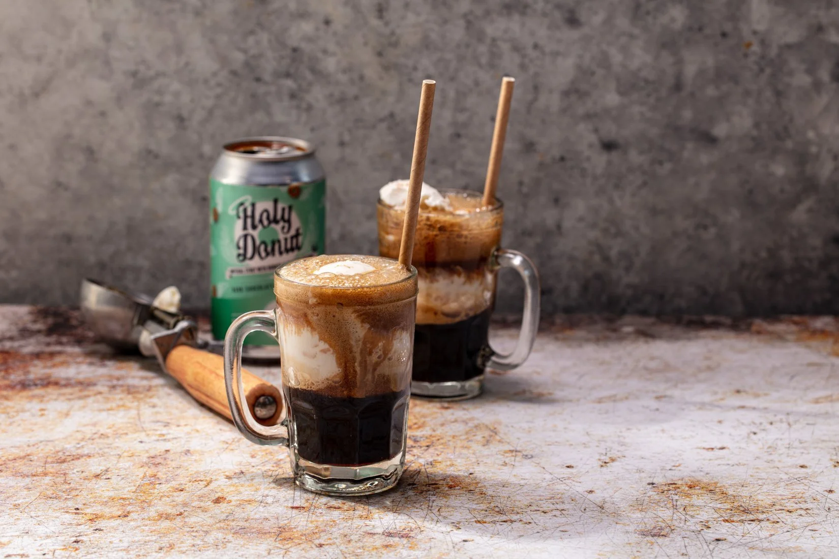 Two glass mugs filled with root beer floats, each with a straw, on a rustic surface, accompanied by a can labeled "Holy Donut" in the background and an ice cream scoop.