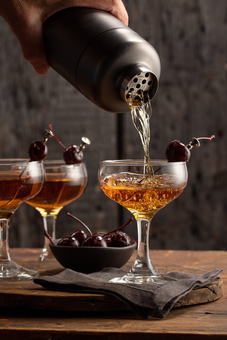 Cocktails being poured from a shaker into coupe glasses garnished with cherries, placed on a wooden table with a bowl of cherries.