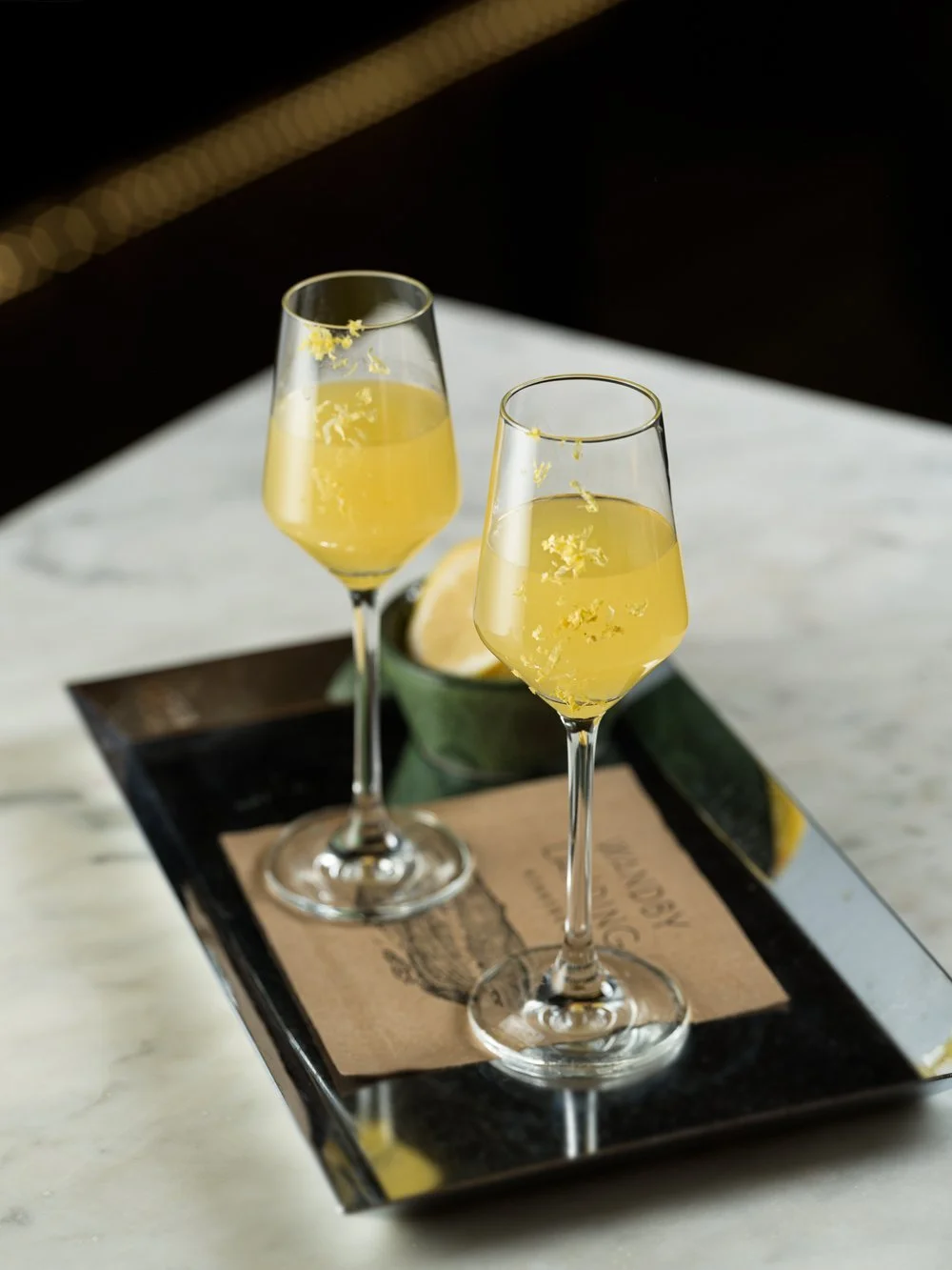 Two yellow cocktails in stemmed glasses with lemon garnish on a tray with a brown napkin.