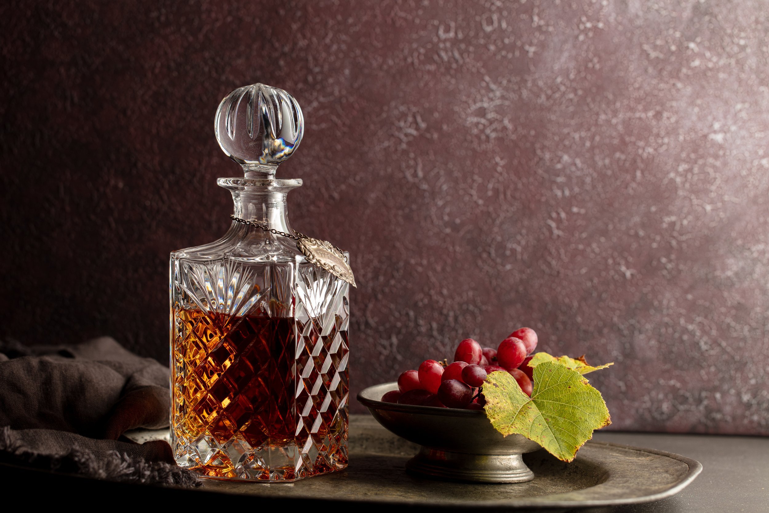 A crystal decanter with brown liquid next to a plate of red grapes and a vine leaf.