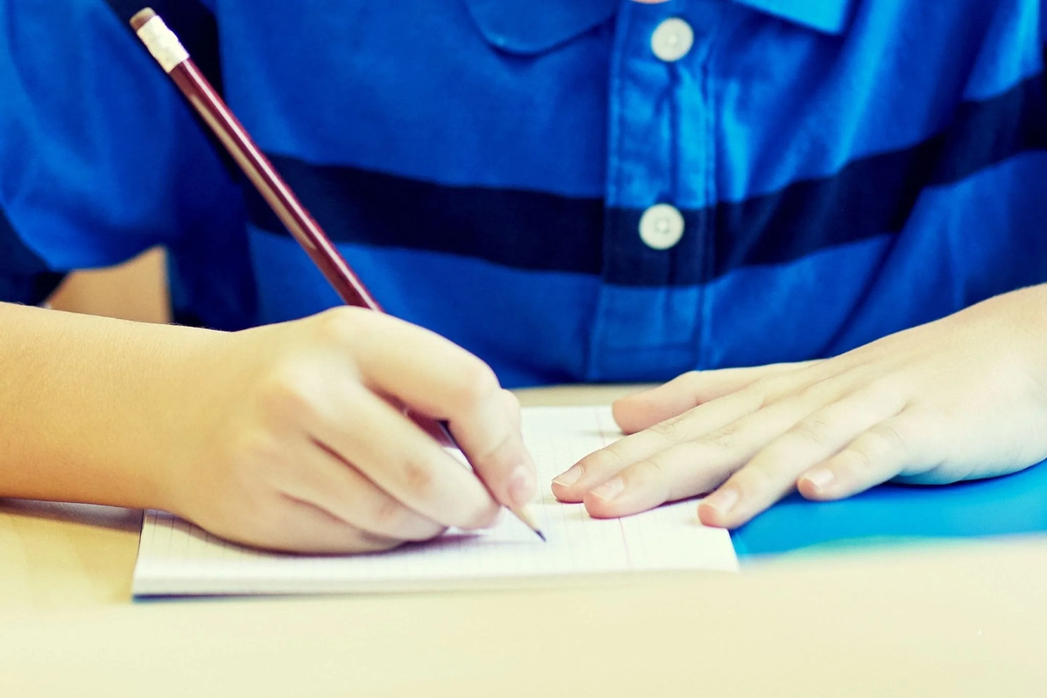 **Alt text:** Young student writing with a pencil in a notebook at a desk.