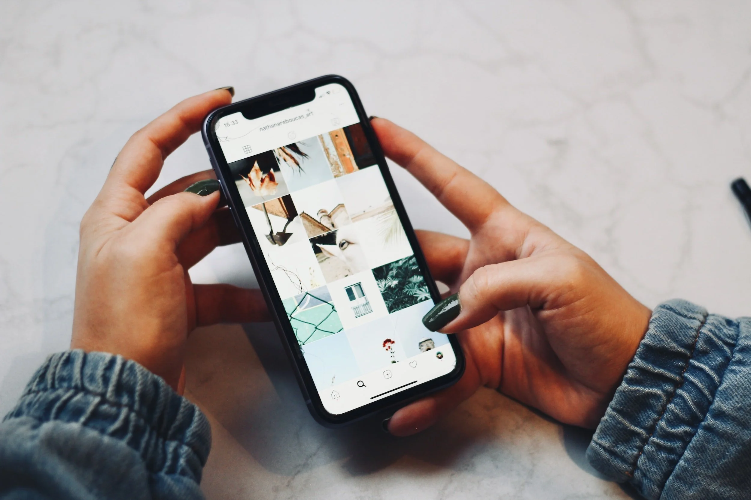 Person holding a smartphone displaying a social media feed with photo thumbnails, wearing a denim jacket.