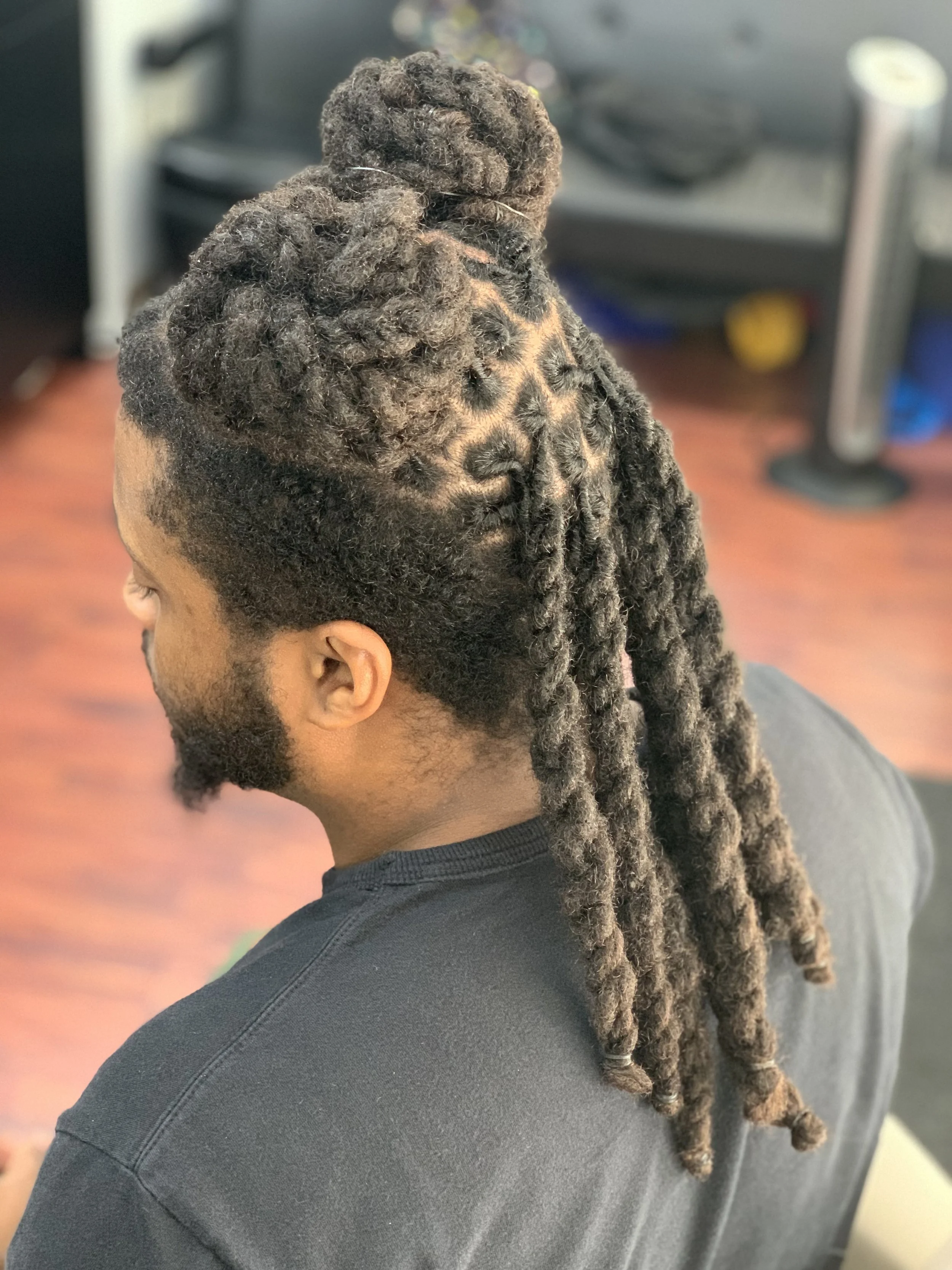 Person with neatly styled dreadlocks, featuring a double bun and long twisted sections, sitting indoors.