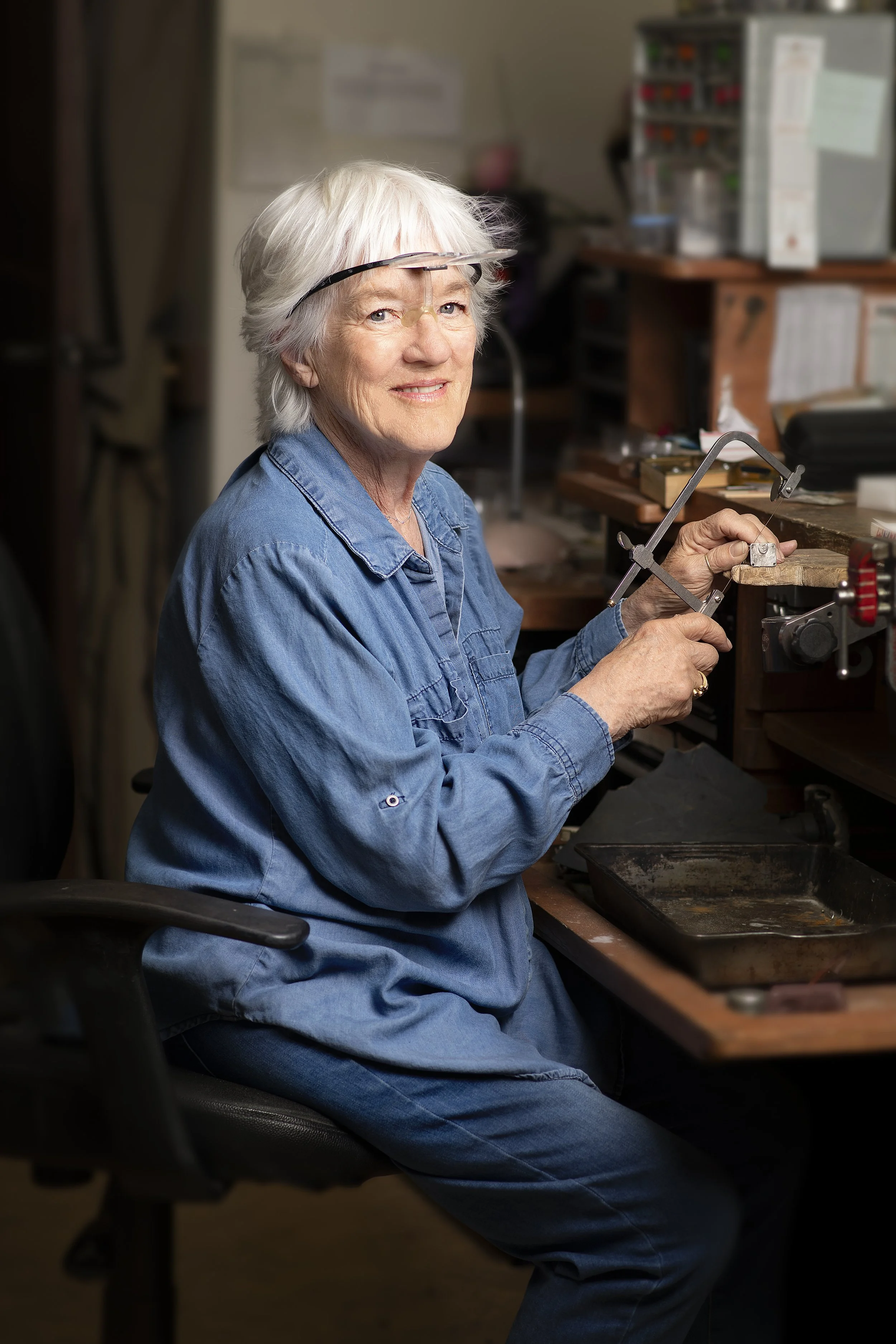 An woman with white hair, wearing a denim shirt, sitting at a workbench in a workshop. She is using a jewelry saw to cut a piece of silver to make a design , with various tools and storage containers in the background.