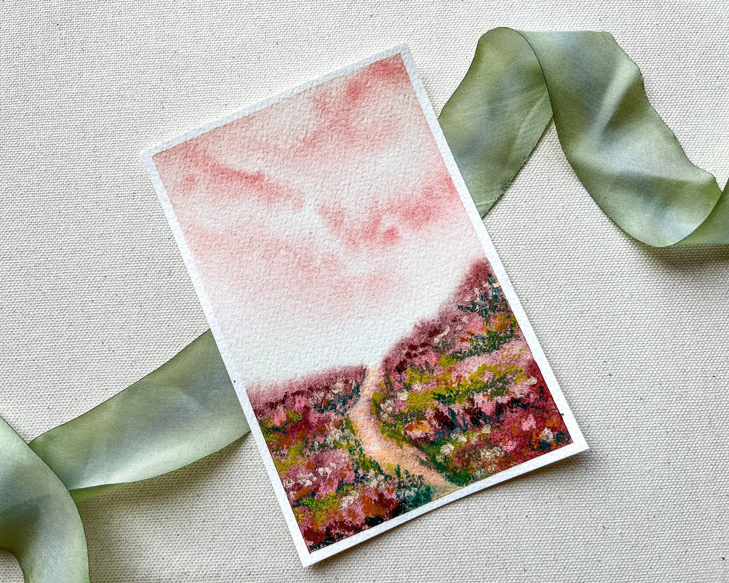 Full view of the pink pastel mixed media landscape with thin white border, atop a green silk ribbon
