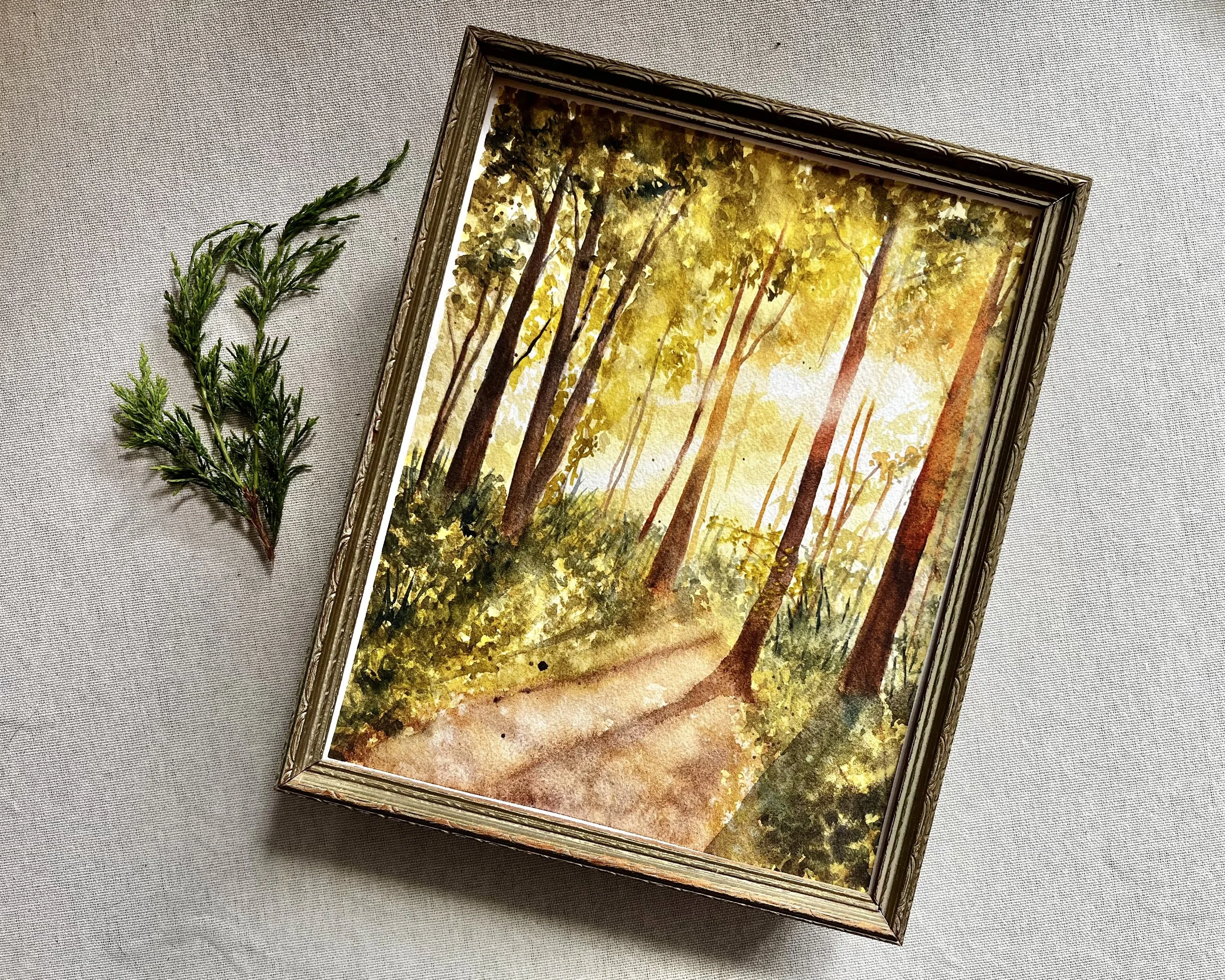 Framed watercolor painting of a dirt path through a forest with sunlight shining down through the leaves