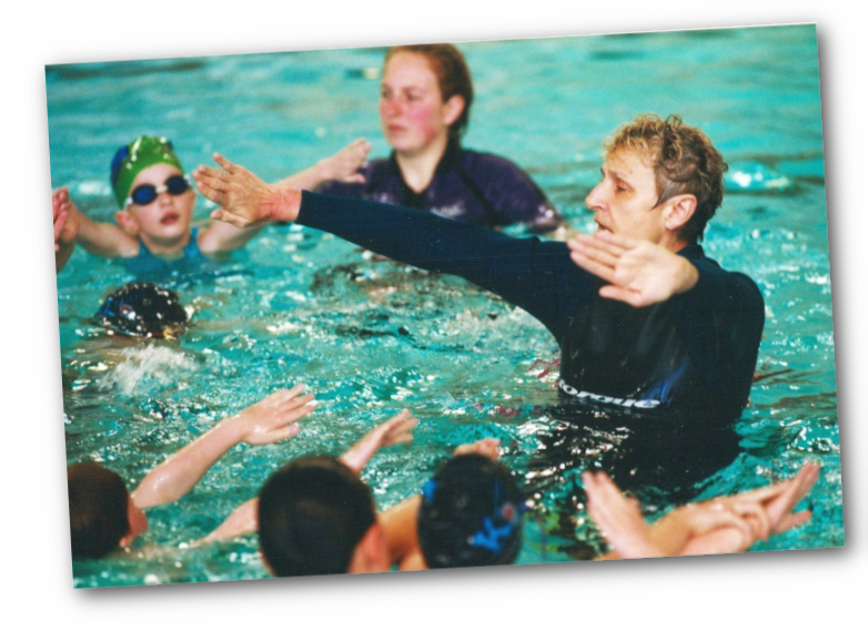 In a swimming pool, Doctor Monika is instructing a group of children, some of whom are reaching out to her, while a girl with a green swim cap and goggles is looking on.