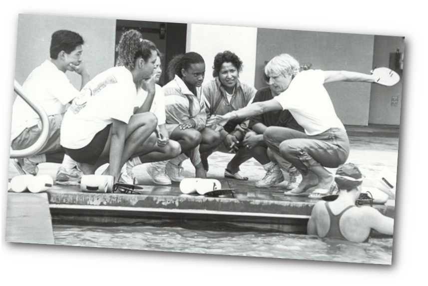 Doctor Monika coaching a diverse group of young female swimmers sitting on the poolside during a training session.