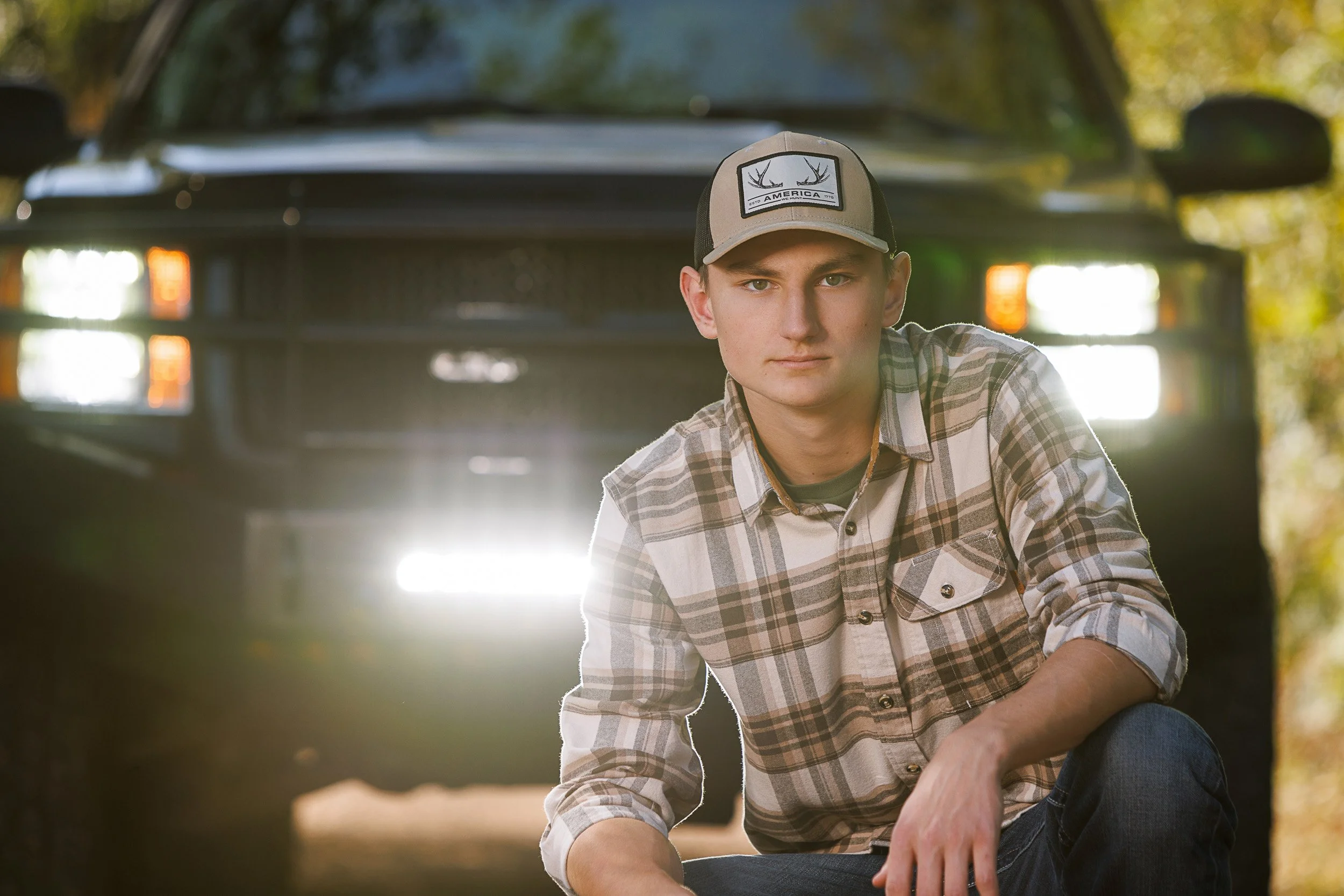 Senior guy wearing a plaid shirt and trucker hat with a black pickup truck with sunlight filtering through trees for a  Ladysmith, WI senior.