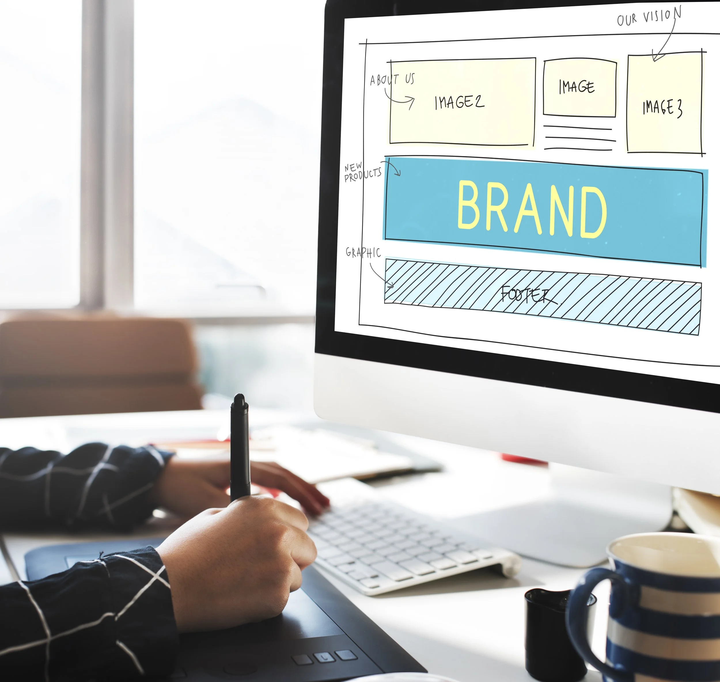 A designer working on a website mockup on a computer, with a hand drawing on a digital tablet. The monitor displays a wireframe layout including sections labeled 'Our Vision,' 'About Us,' images, 'Brand,' 'Graphic,' and 'Footer.' A coffee mug and a drawing tablet are on the desk.