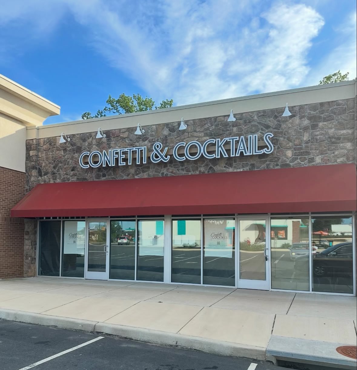 Exterior of a restaurant called Confetti & Cocktails with a stone facade, red awning, and glass doors, situated in a shopping plaza area under a blue sky with clouds.