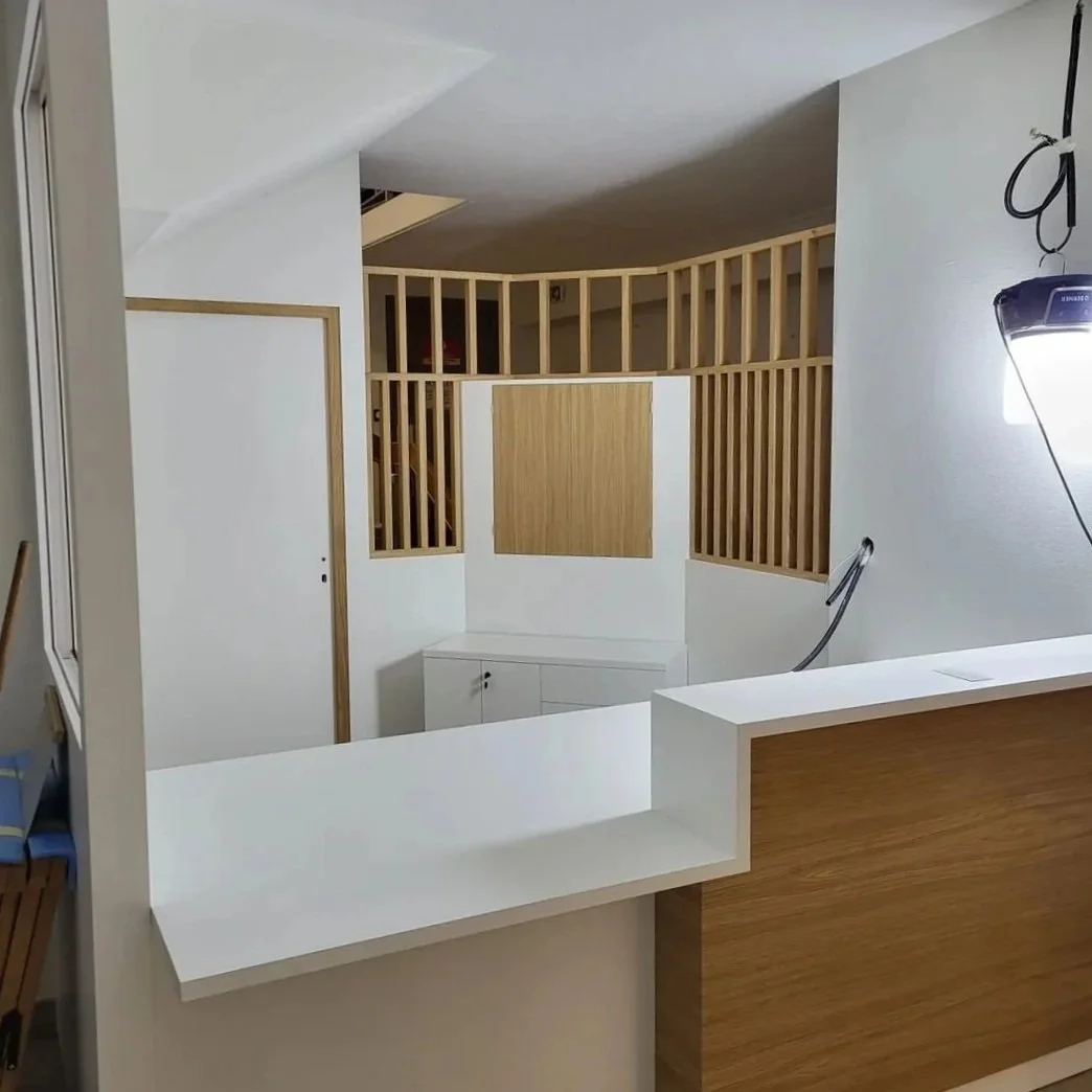 Intérieur d'un espace en cours de rénovation avec un comptoir blanc en premier plan, des murs en blanc et des éléments en bois clair, notamment un panneau mural et une structure en bois autour d'une zone partiellement séparée.