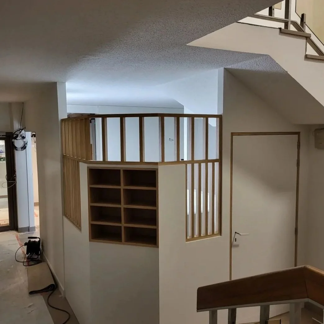 Intérieur d'une maison en cours de rénovation avec une mezzanine en bois, des escaliers en bois avec rampe métallique, et des murs blancs.