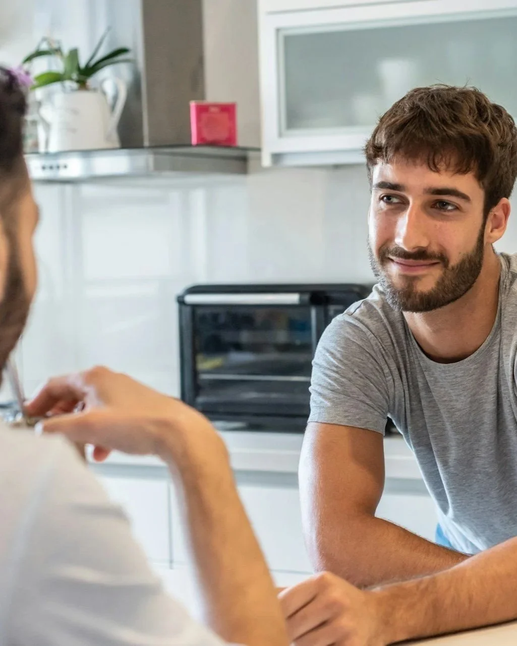 Two men having a conversation in a kitchen, one facing the camera and smiling, the other with his back to the camera.