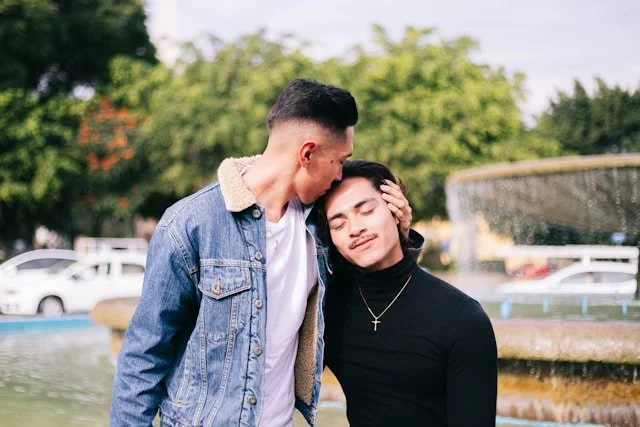 A gay couple standing close together outdoors by a fountain, one gently holding the other's head, with trees and cars in the background.