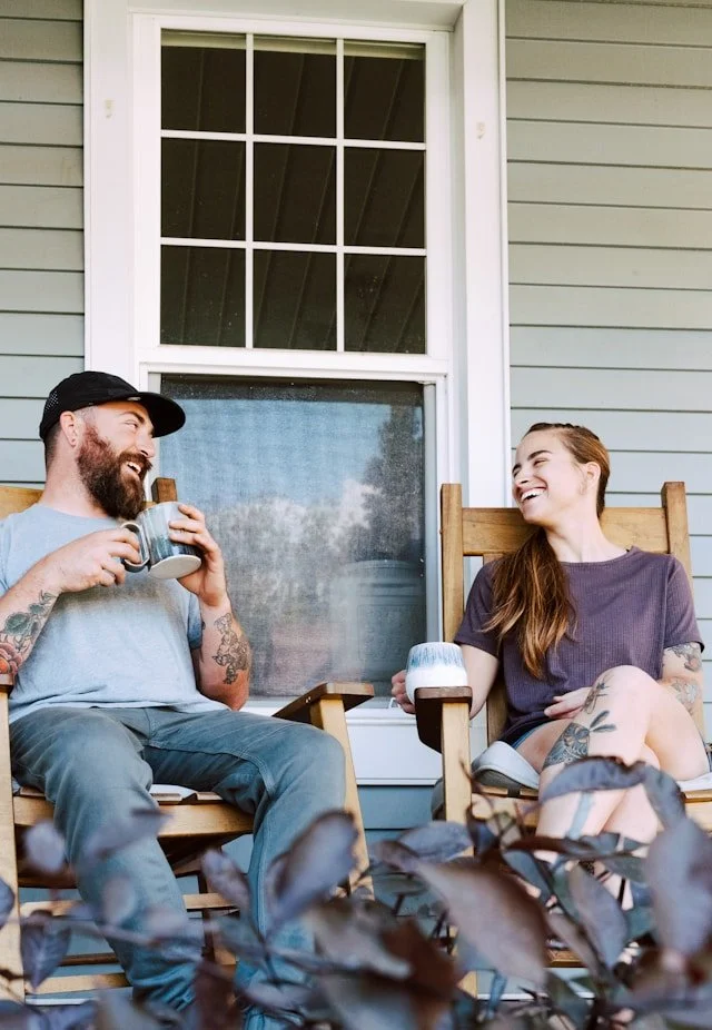A man and woman sitting on a porch in wooden chairs, enjoying drinks and smiling at each other.