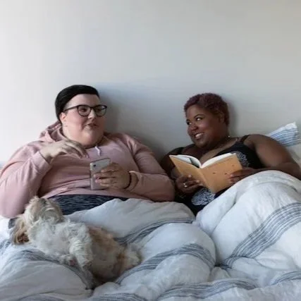 Two women sitting on a bed with a dog lying between them, one woman holding a phone and the other reading a book.