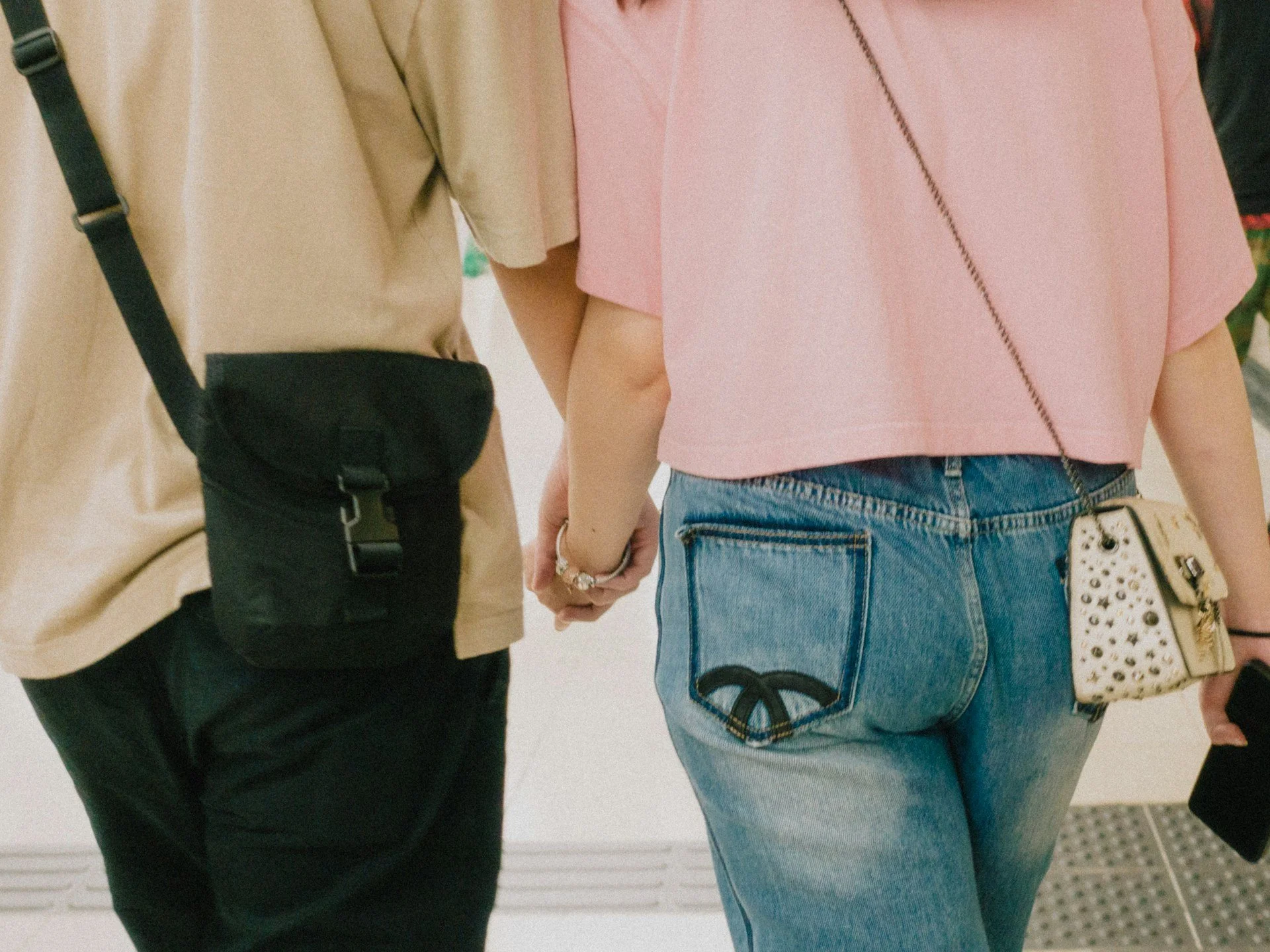 Couple holding hands, one wearing a beige shirt with a black bag, and the other wearing a pink top with a leopard print purse, both in casual jeans.