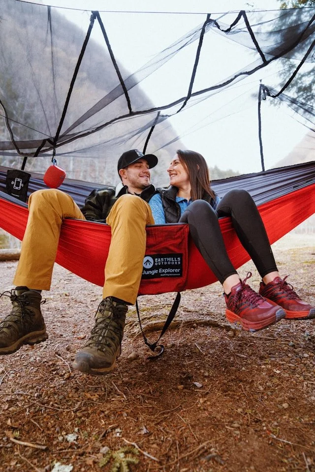 A smiling couple sitting together in a red hammock under a tarp, surrounded by trees and outdoor scenery.