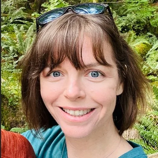A woman with shoulder-length brown hair, wearing sunglasses on her head, smiling outdoors in a green, leafy setting.