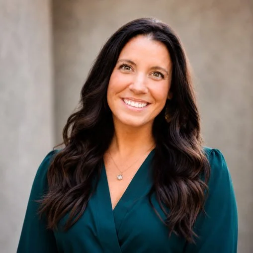 A woman with long dark hair smiling, wearing a teal blouse with a v-neck and a necklace, standing outdoors with a neutral background.