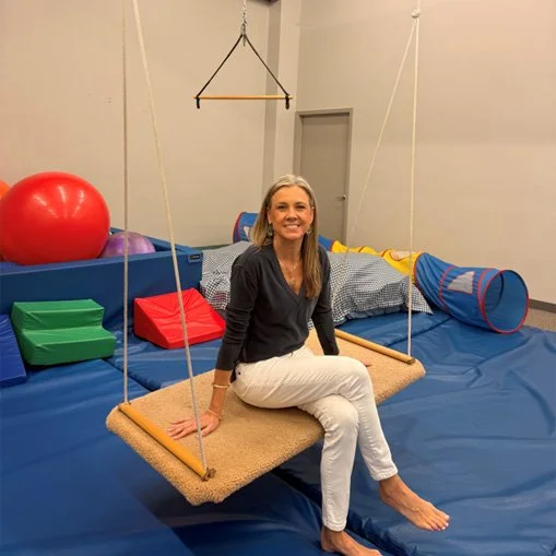 A woman sitting on a therapy swing in a room with exercise equipment like a large red exercise ball, foam blocks, a tunnel, and a padded floor.